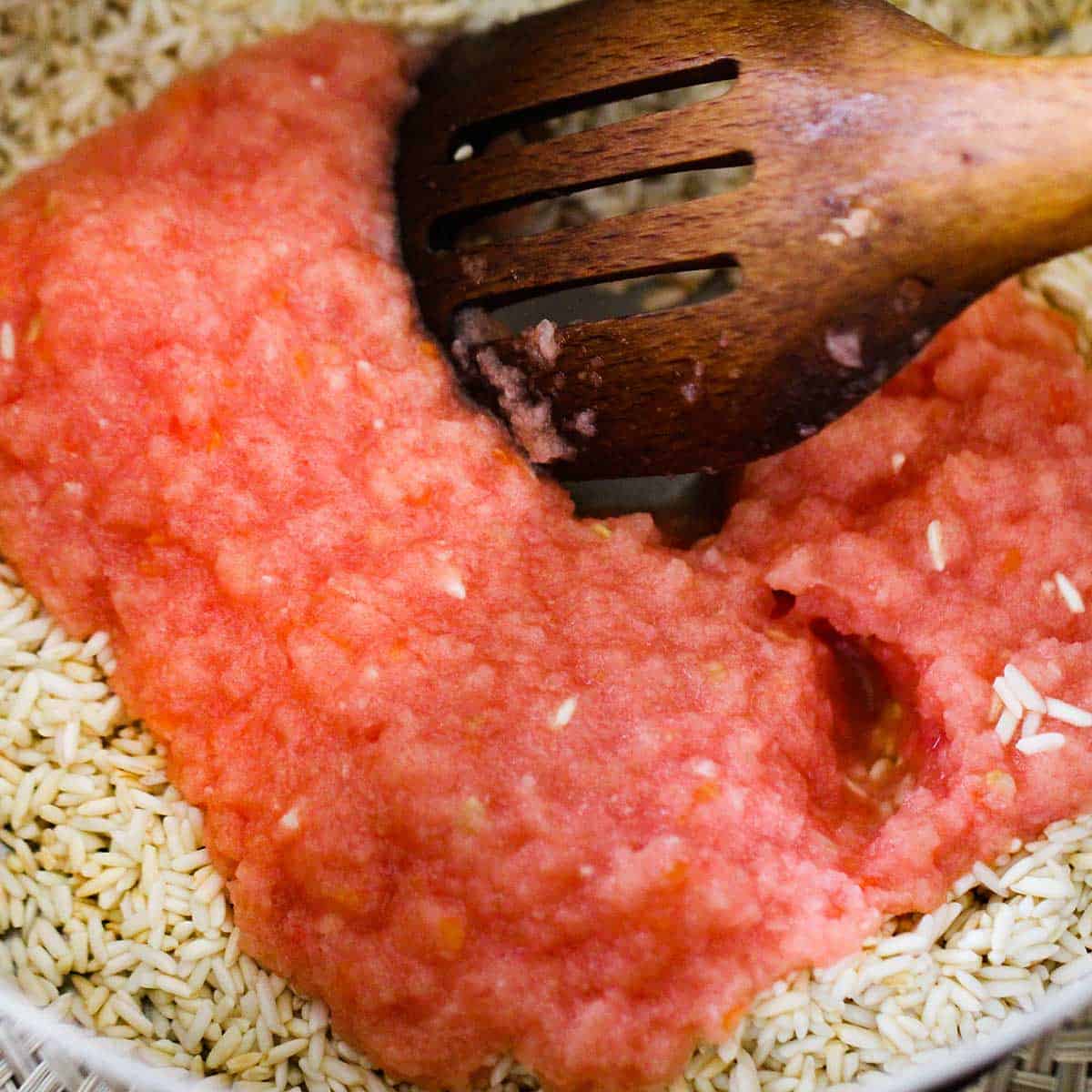 A wooden slotted spoon stirring a puréed tomato and onion mixture into a saucepan filled with toasted long-grain rice.