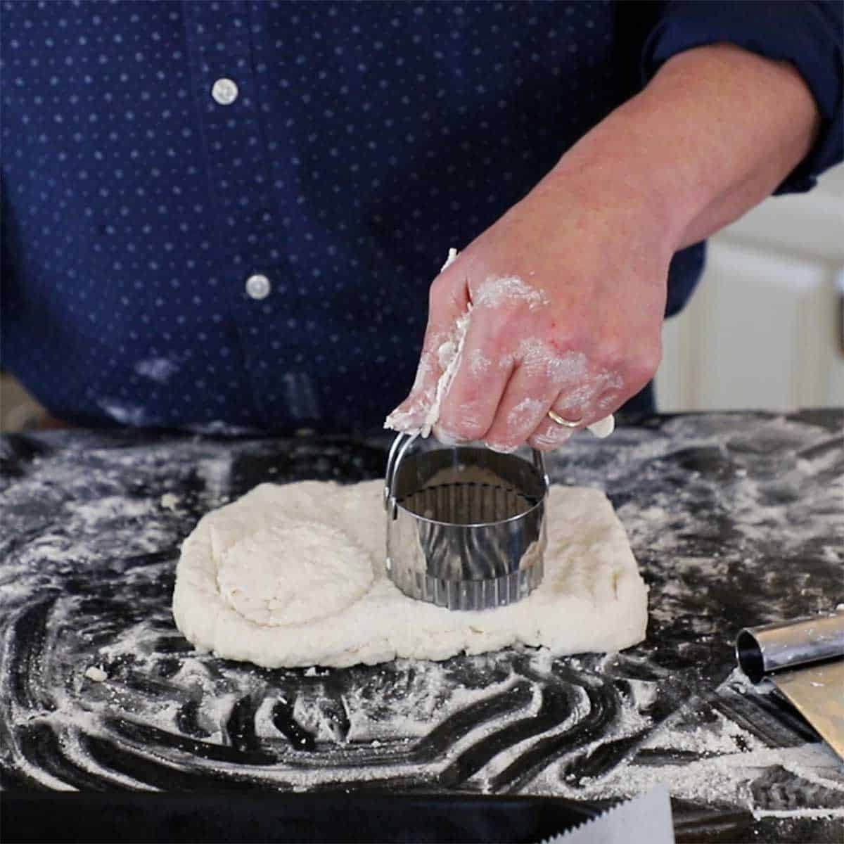 A person using a metal cookie cutter to cut into a rectangle square of biscuit dough on a black marble counter that has been lightly floured.