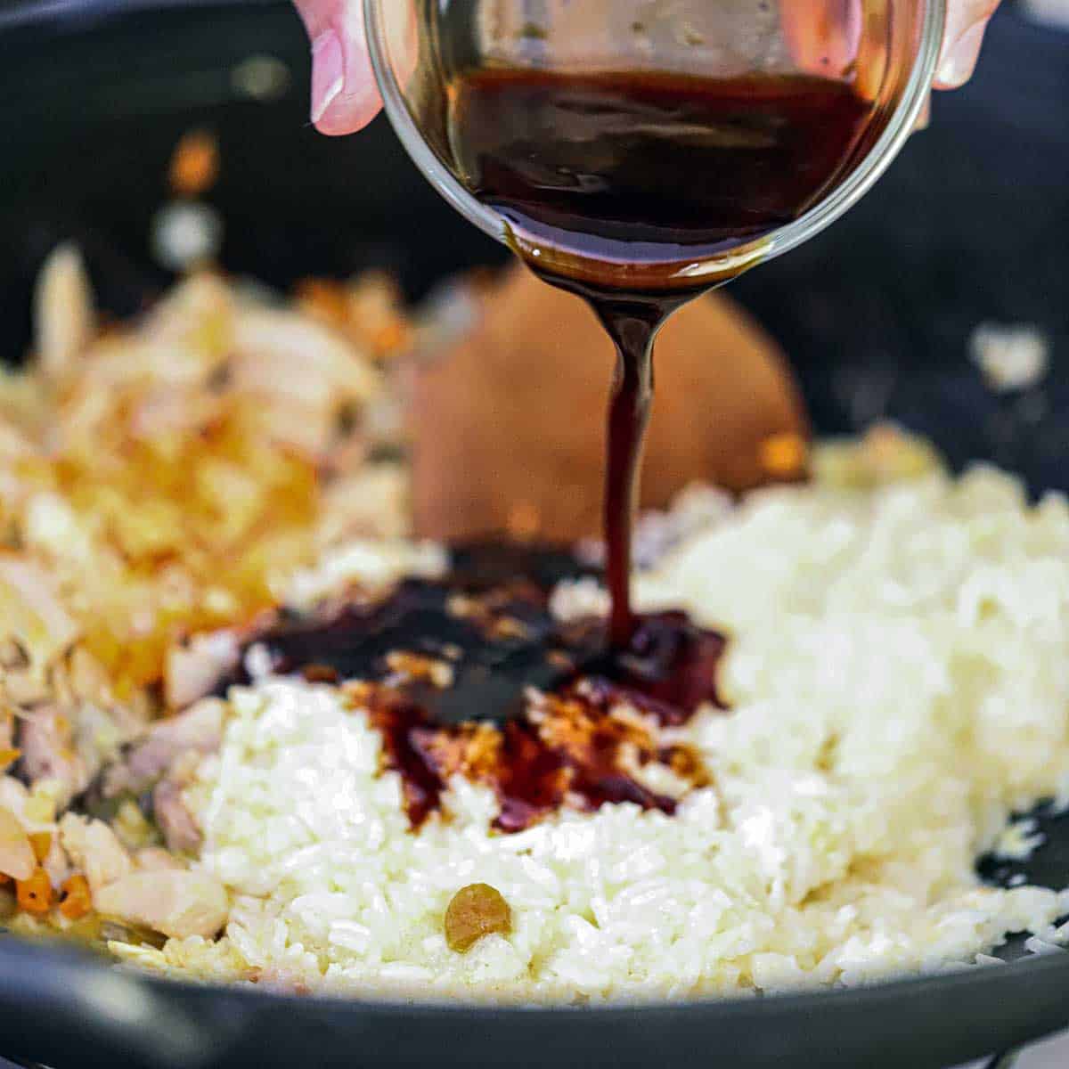 A person pouring a brown Asian sauce over an rice and egg mixture in a wok also with sautéed vegetables and chicken in it.
