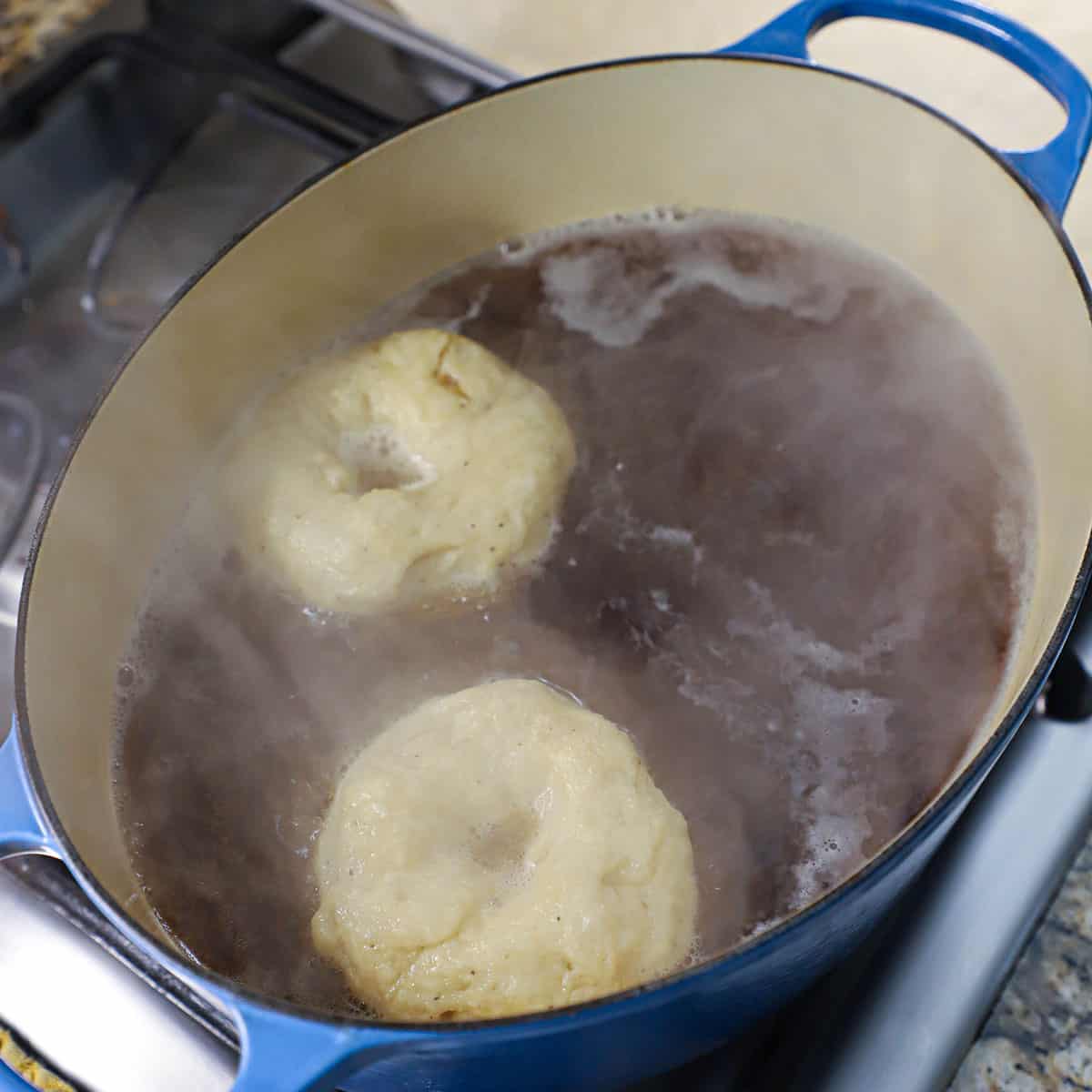 Two bagels boiling in water with molasses in an oval Dutch oven on a stove.