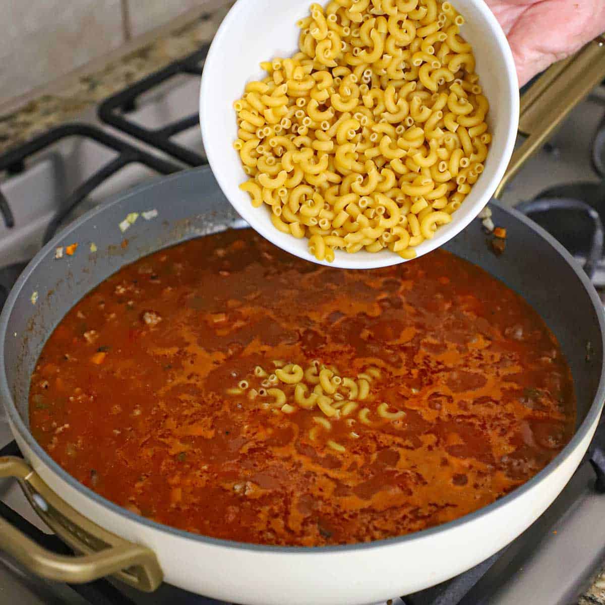 A person dumping uncooked macaroni from a large white bowl into a skillet filled with a simmering tomato and beef sauce.