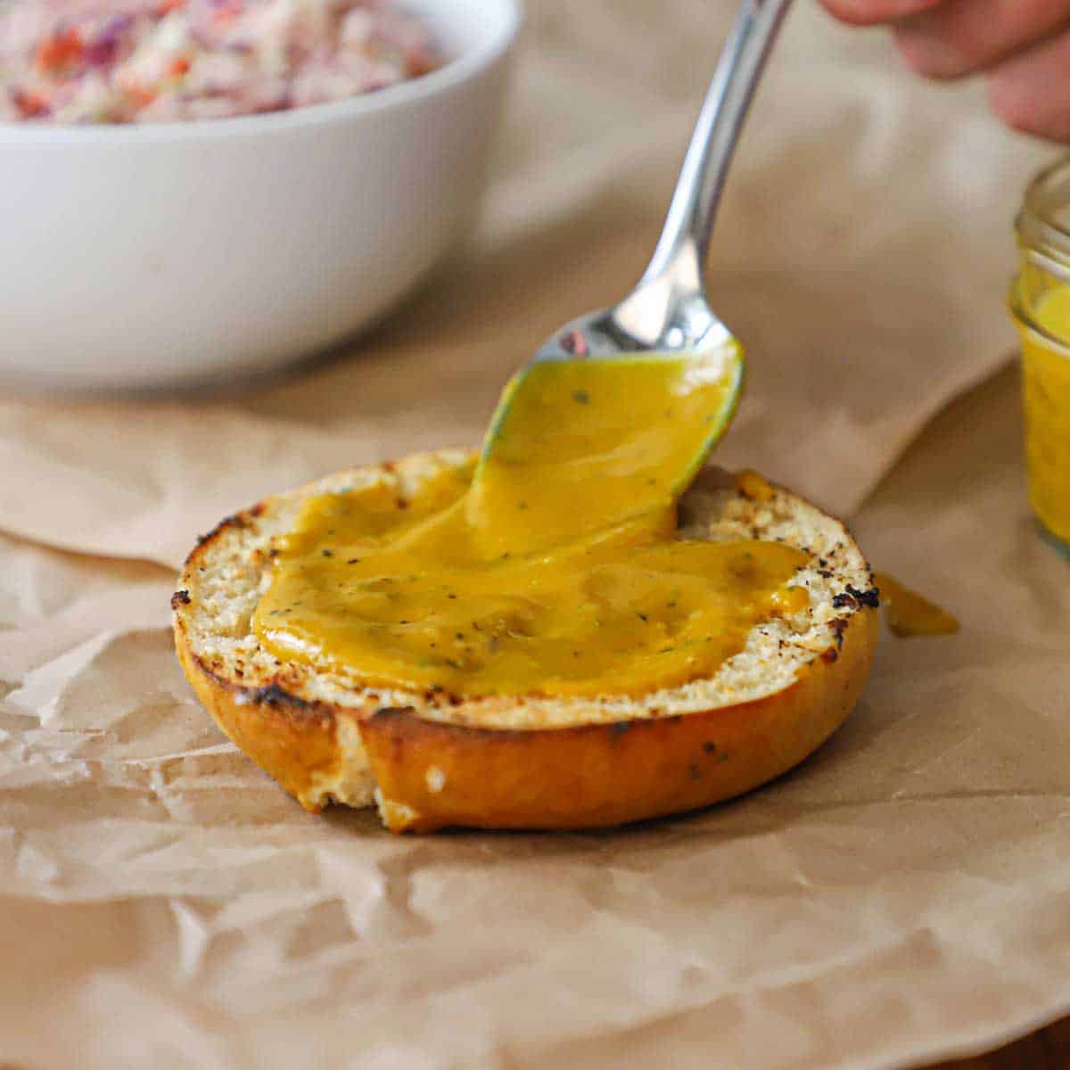 A person using a spoon to smear a Carolina mustard sauce over the bottom half of a toasted pretzel bun.