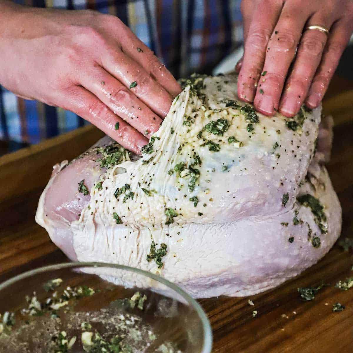 A person rubbing a butter and herb mixture under the skin of an uncooked whole turkey breast on a wooden cutting board.