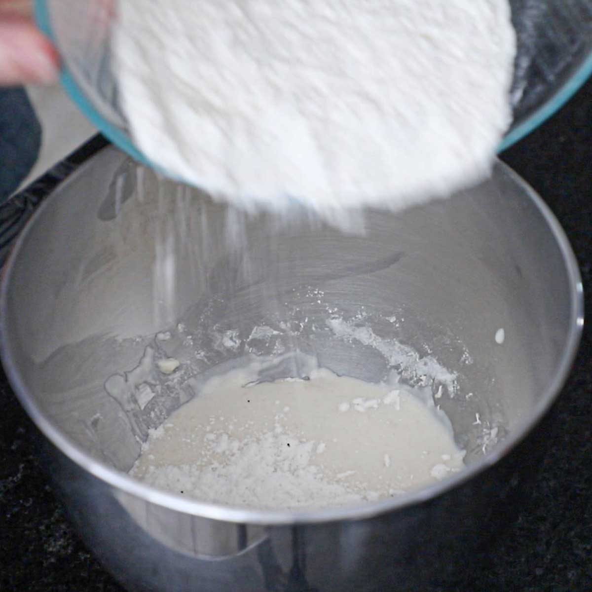 A person adding a flour and yeast mixture into the bowl of a mixer that contains a bagel dough sponge.