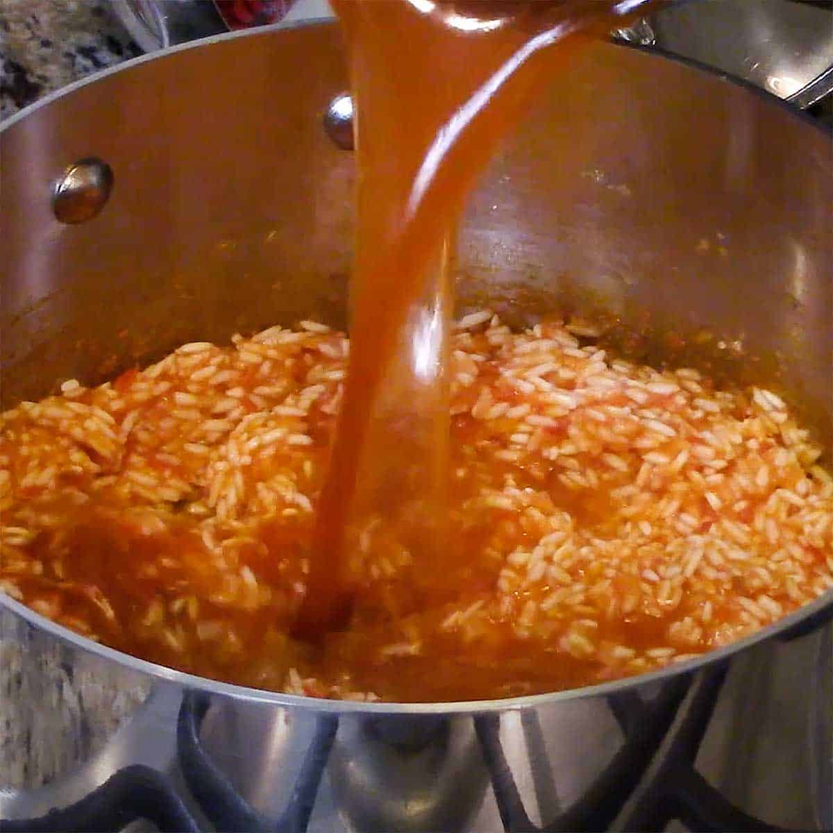 A person pouring a seasoned chicken stock from one pan into another pan filled with long-grain rice and puréed tomatoes and onions.