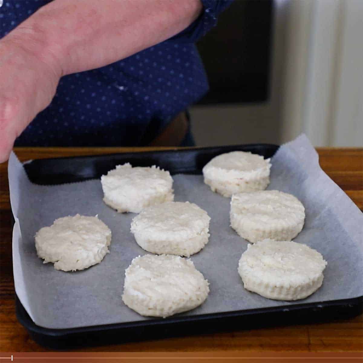 A person placing freshly cut rounds of buttermilk biscuit dough on a square baking dish that has been lined with parchment paper.
