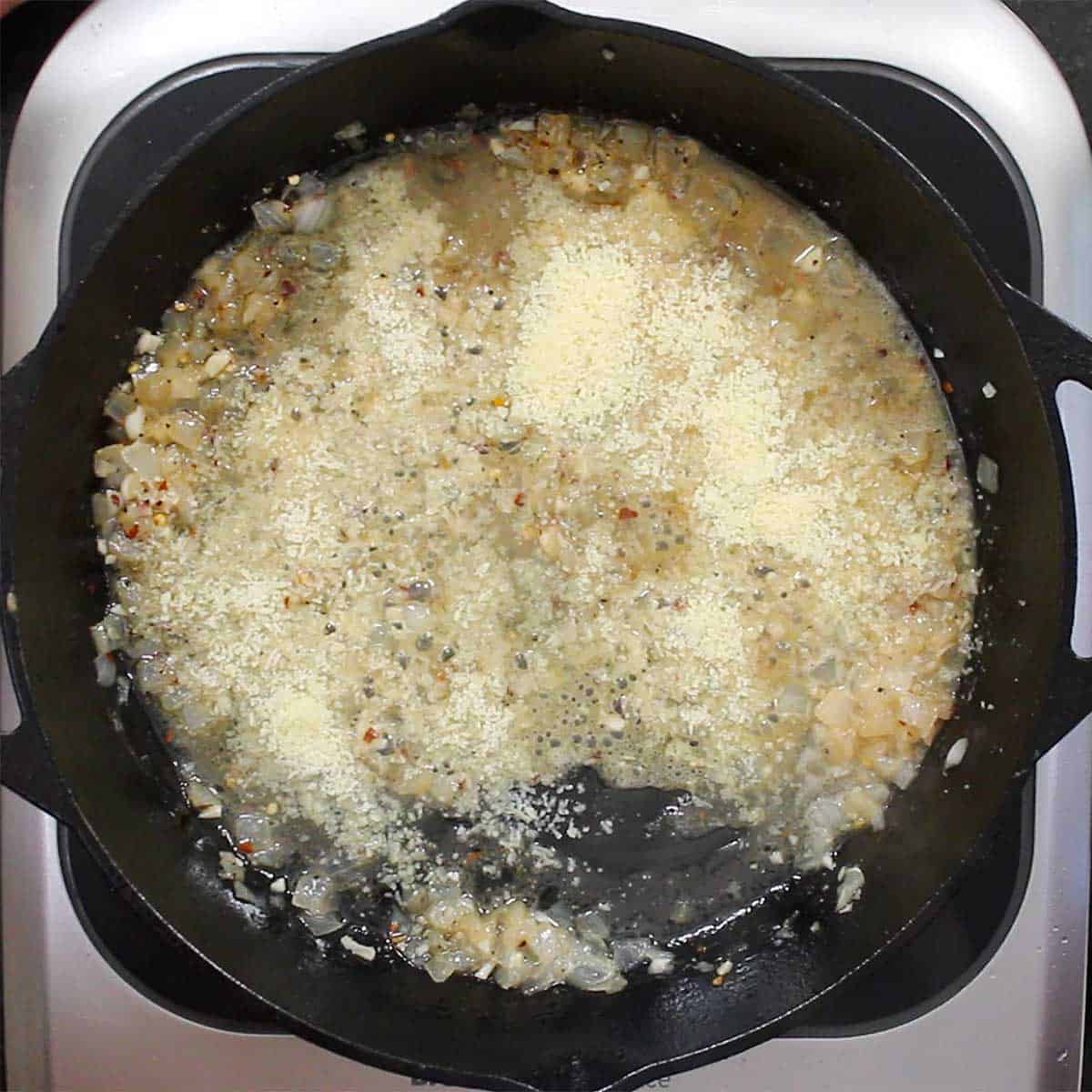 A large black cast-iron skillet filled with sautéed onion and garlic and is simmering in white wine, broth, and Parmesan cheese.