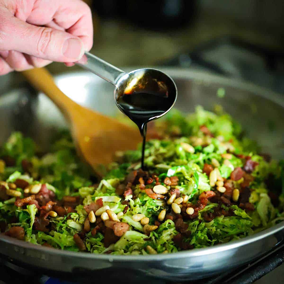 A person pour balsamic vinegar from a tablespoon into a skillet filled with sautéed shaved Brussels, crispy pancetta, and pine nuts.