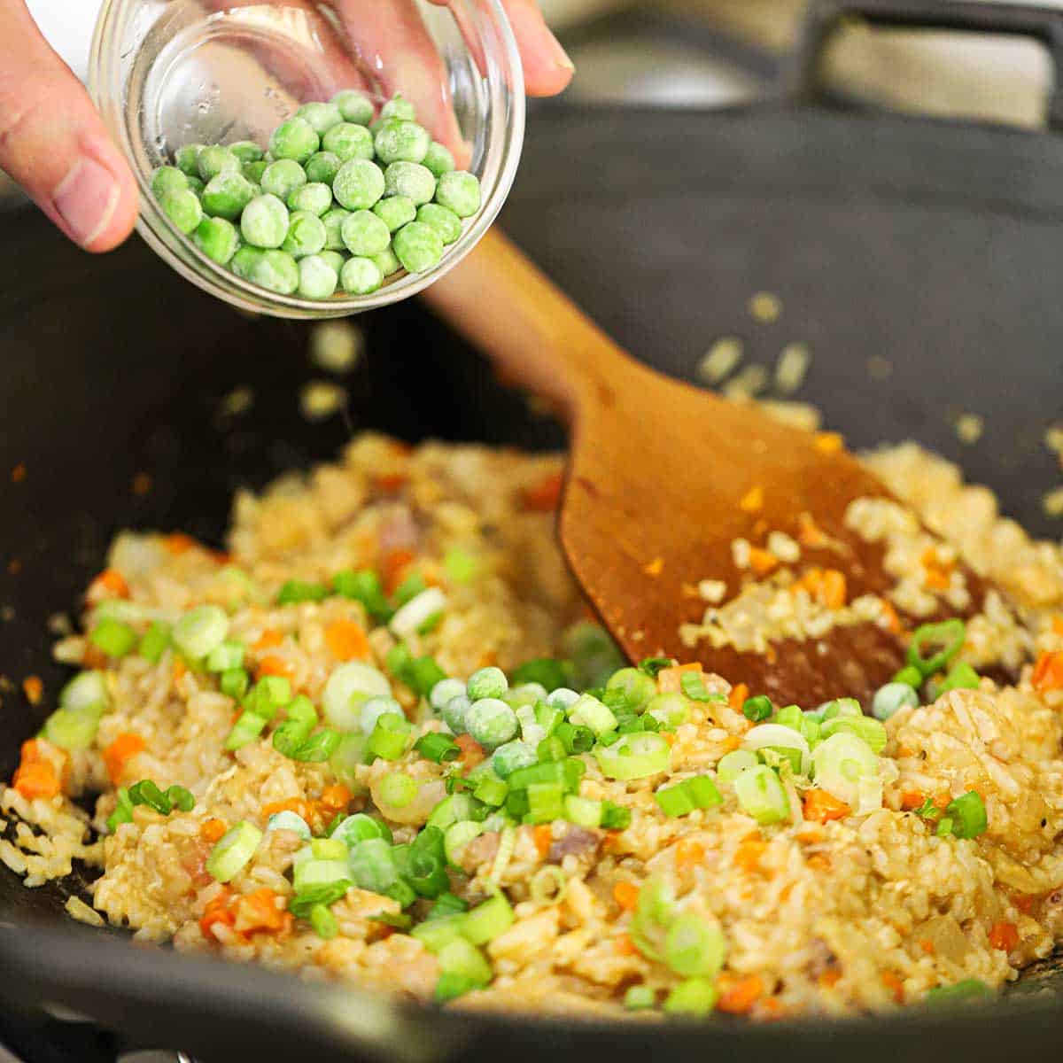 A person transferring frozen peas from a small glass bowl into a wok filled with chicken fried rice with a large wooden spatula resting in the middle of the rice.