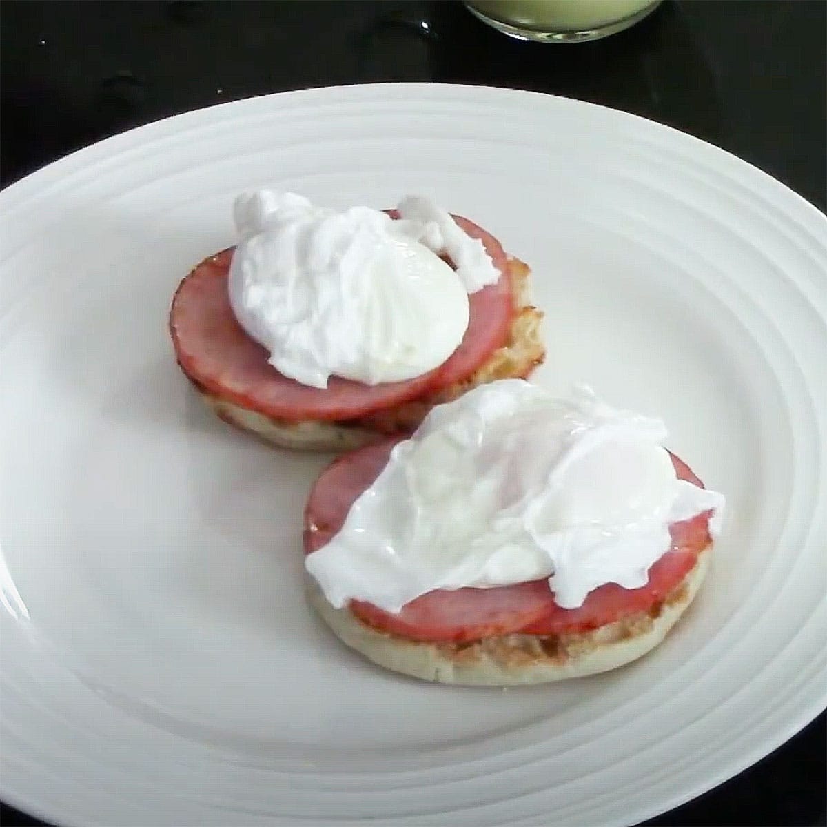 A white dinner plate filled with a pair toasted English muffins each topped with crispy Canadian bacon slices and a poached egg.