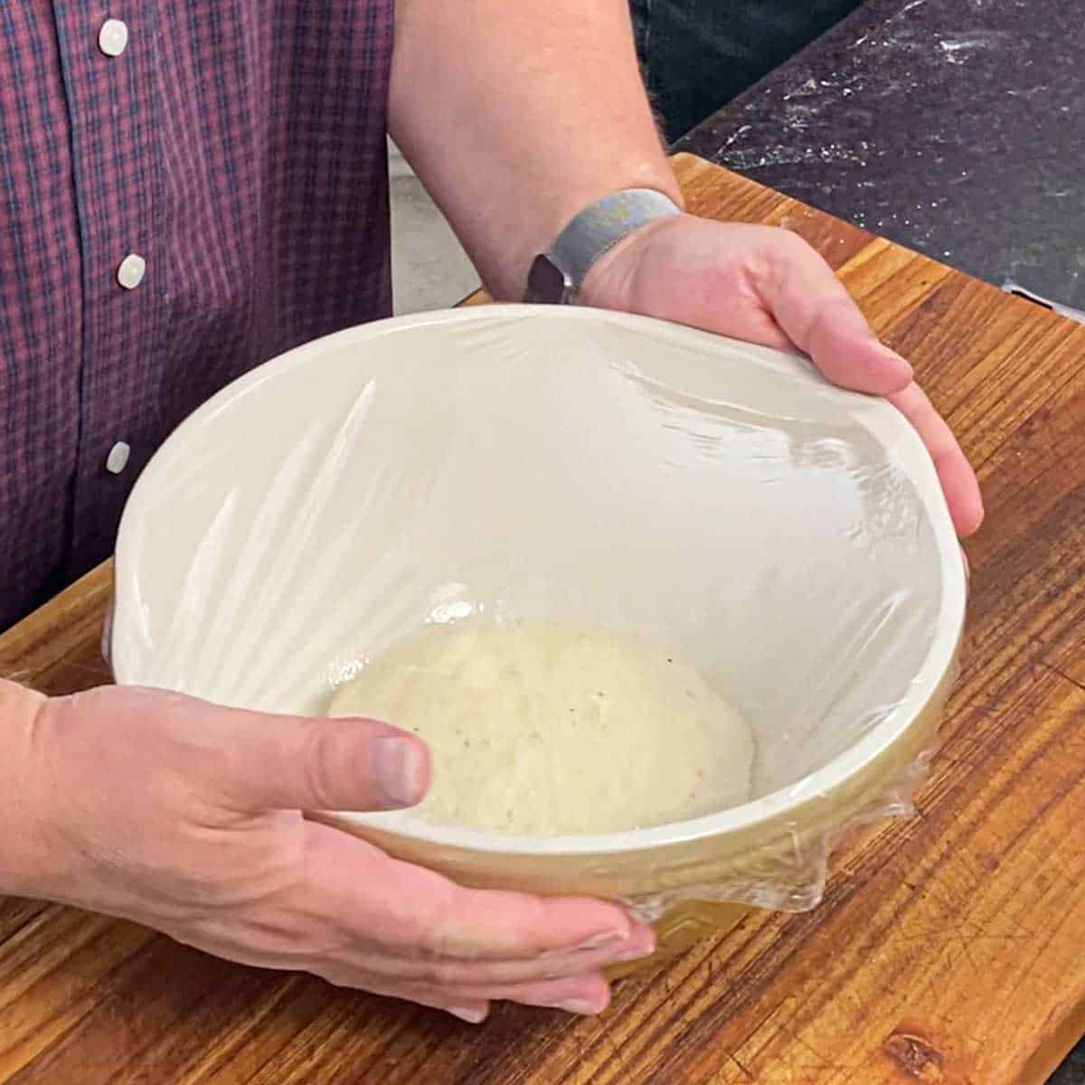 A person holding the sides of a ceramic bowl that has bagel dough in it and is covered with clear plastic wrap.