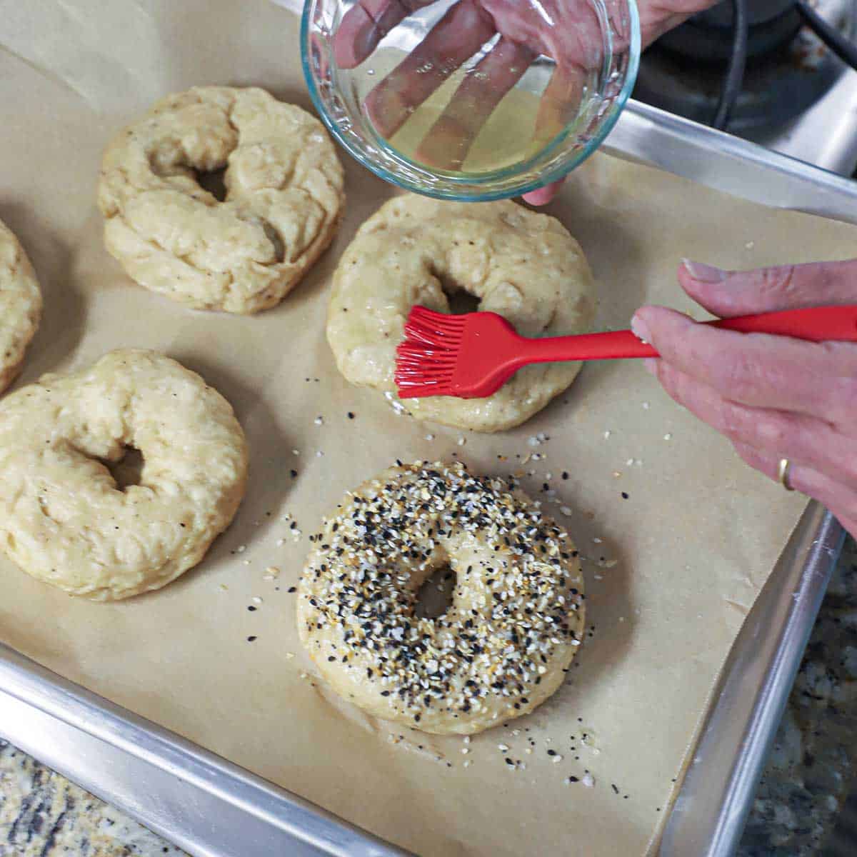 A person using a pastry brush to add an egg white wash over the top of an un-baked bagel sitting next to a bagel that has everything bagel seasoning sprinkled on top.