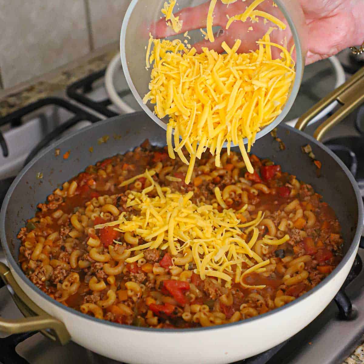 A person dumping grated cheddar cheese from a glass bowl into a large skillet filled with beef chili and cooked macaroni.