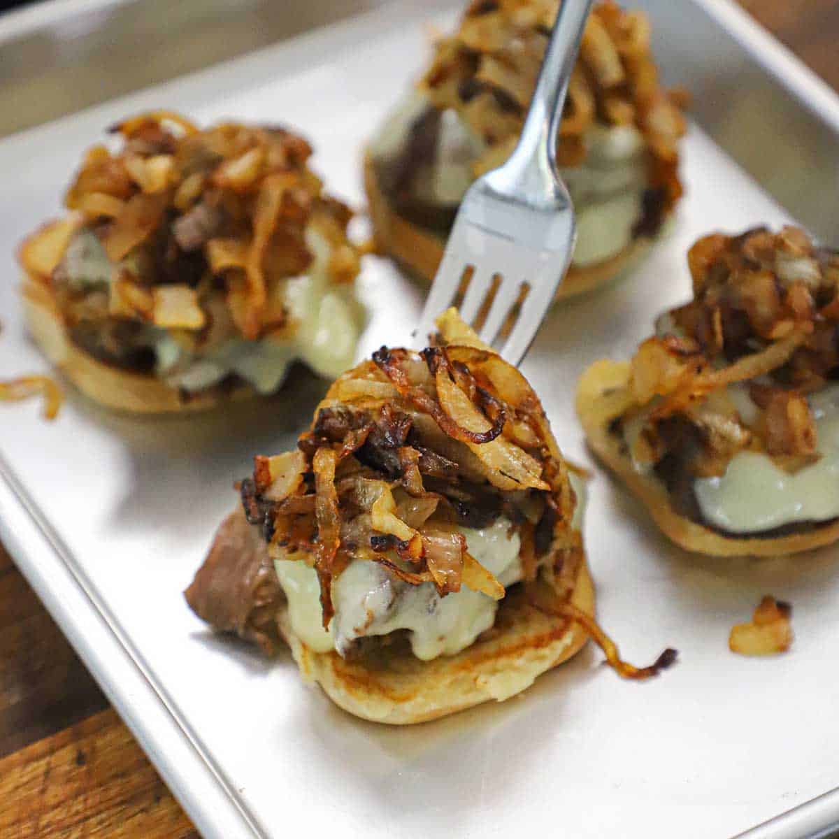 A person using a fork to place caramelized onions on top of a pot roast slider that has melty Swiss cheese on top of the meat.