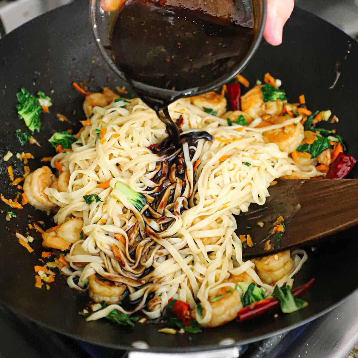 A person pouring a dark Chinese lo mein sauce into a wok that is filled with sautéed vegetables, shrimp, and cooked Asian egg noodles.