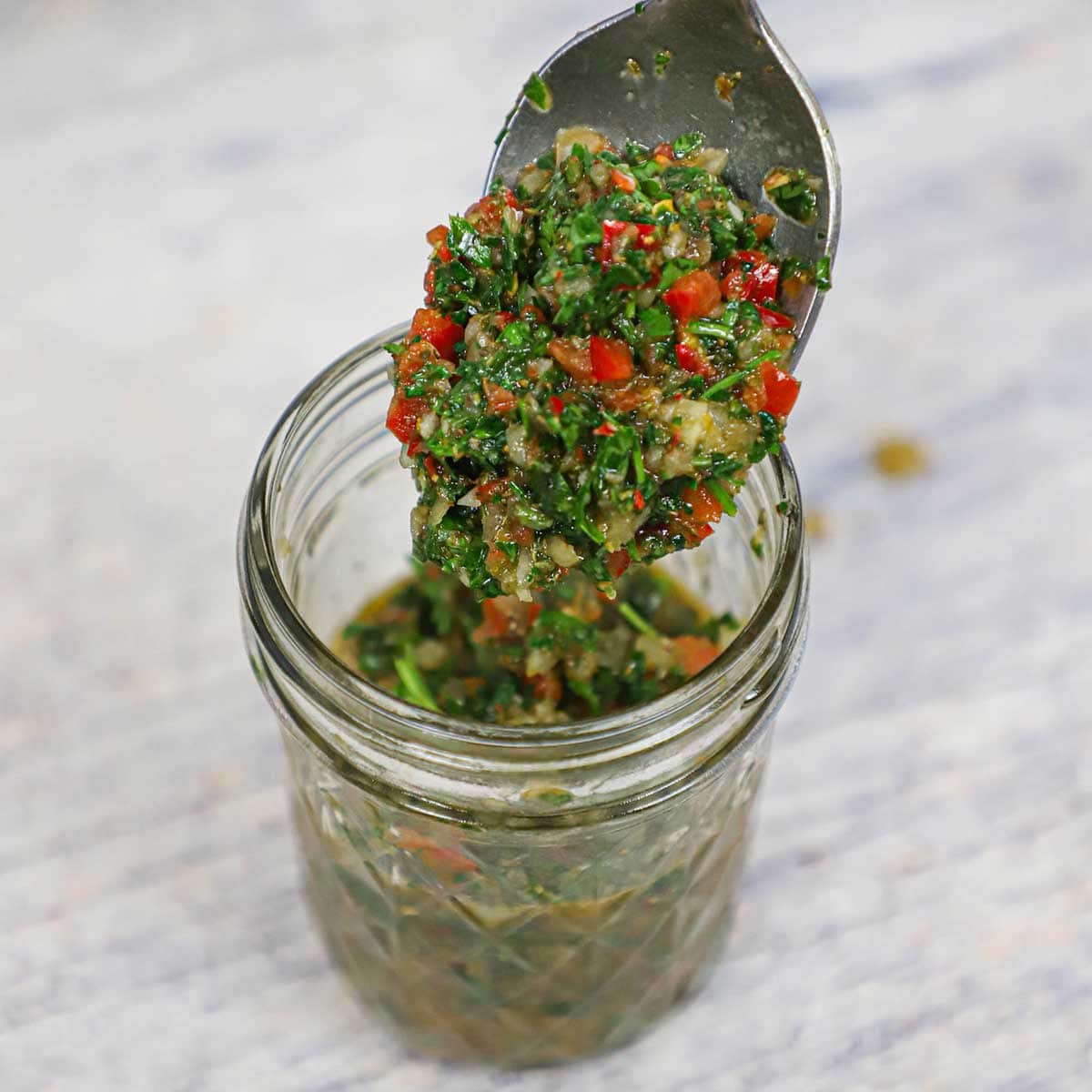 A person holding spoon full of authentic chimichurri sauce over a small glass jar filled with the same.