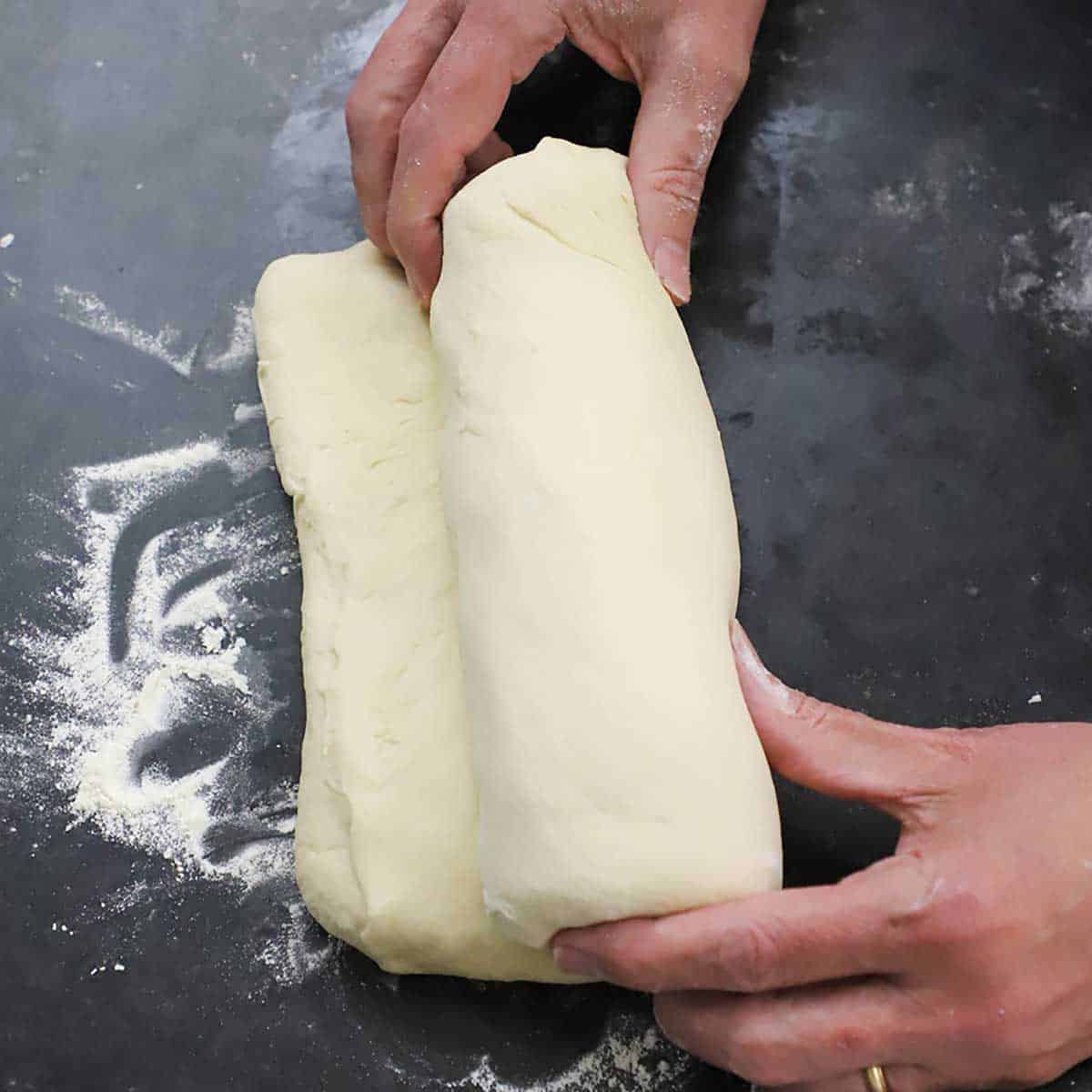 A person rolling a rectangular piece of bread dough into a tight cylinder on a black marble counter top that has been lightly floured.