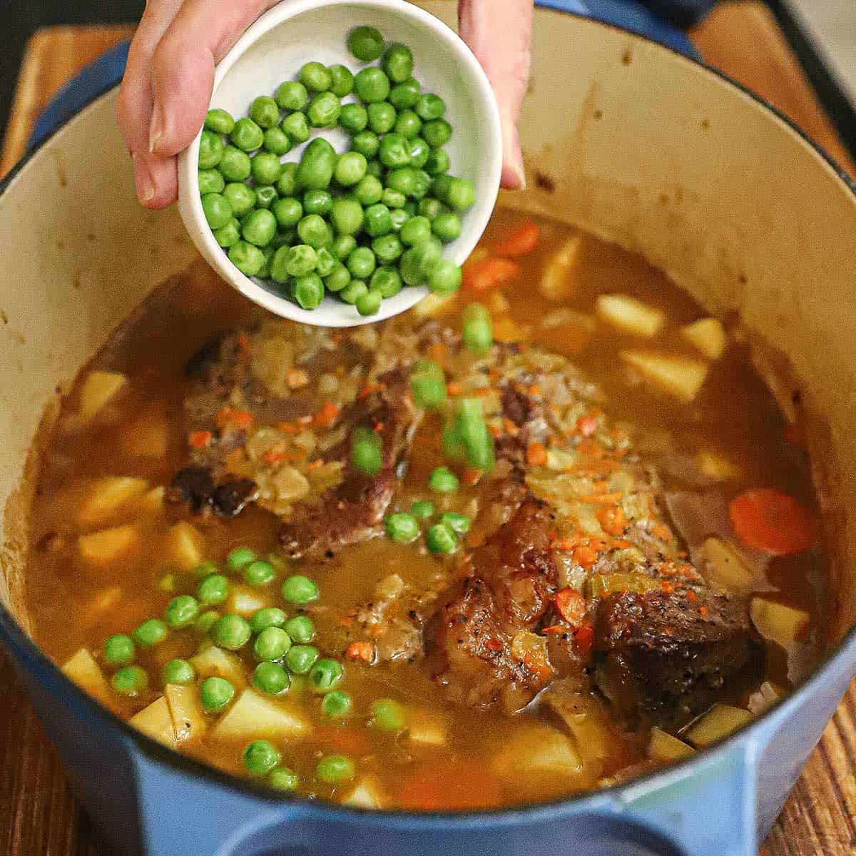 A person transferring sweet green peas from a small white bowl into a Dutch oven that is filled with a classic pot roast, simmering broth, and tender chunks of potatoes and carrots.