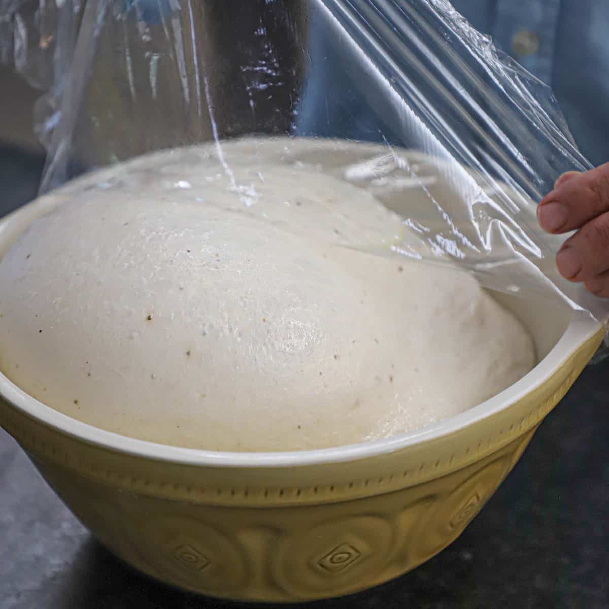 A person removing clear plastic wrap from the top of a ceramic bowl that is filled with bagel dough that has fully risen to the top of the bowl.