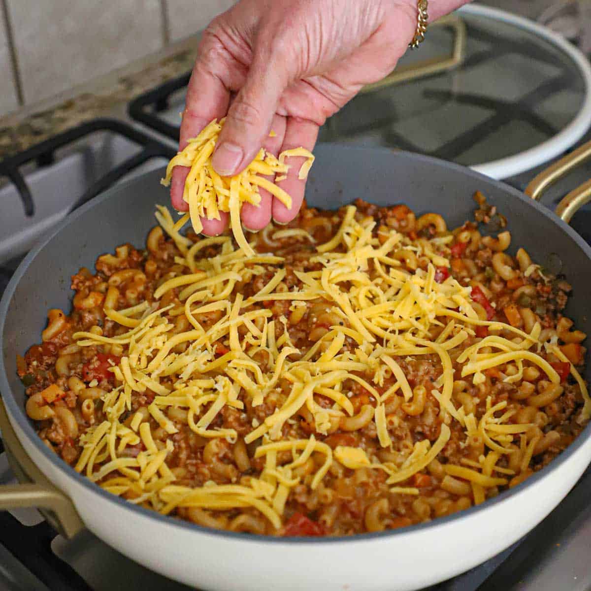 A person sprinkling shredded cheddar cheese over the surface of chili mac in a large skillet.