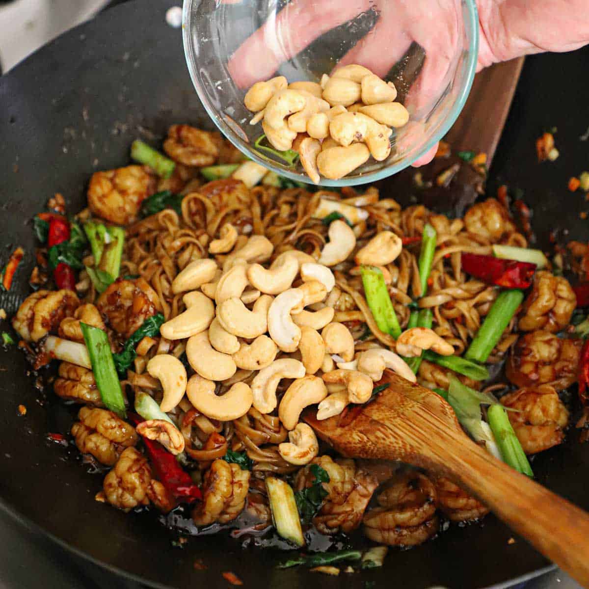 A person dumping roasted cashews from a small glass bowl into a wok filled with freshly prepared shrimp lo mein.