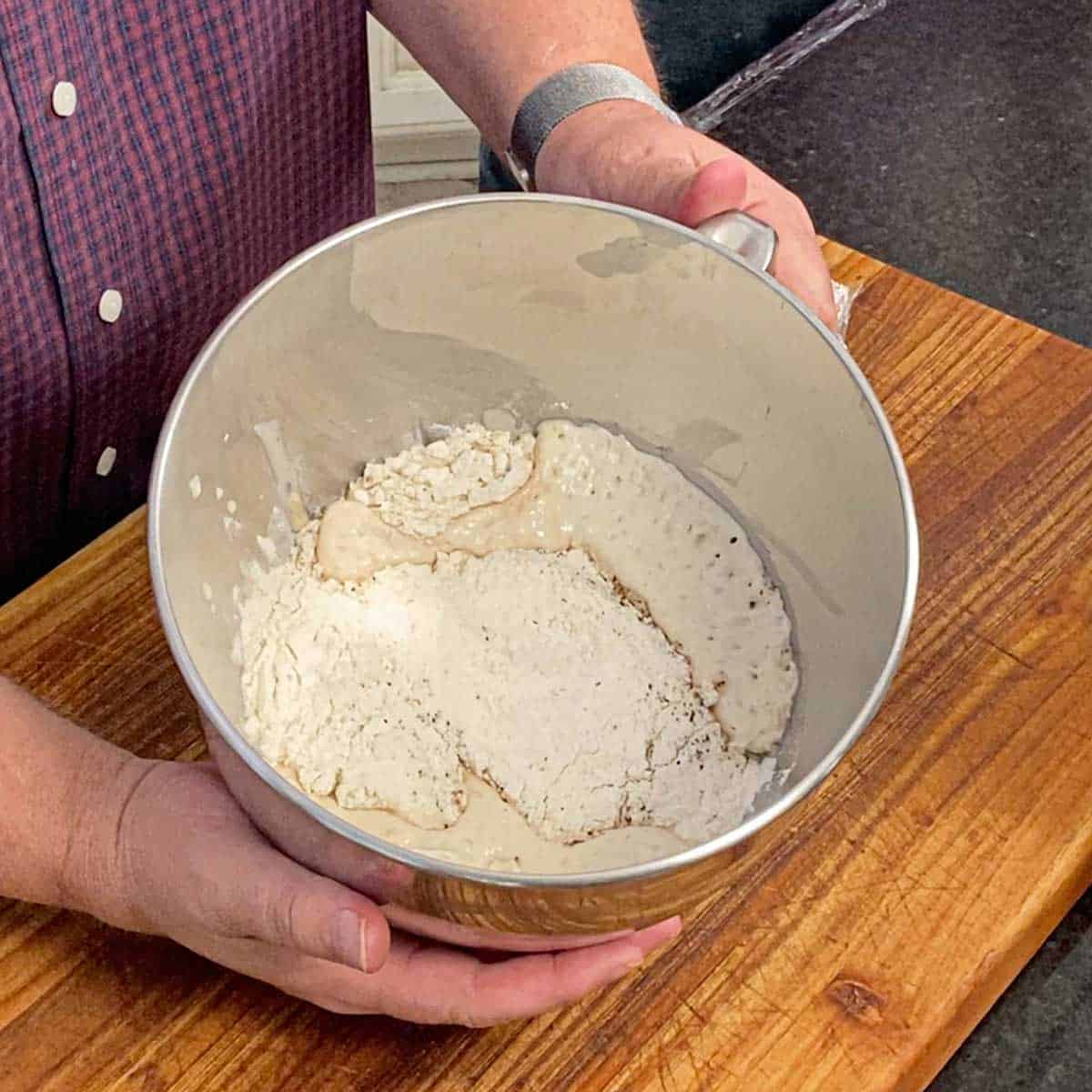 A person holding the bowl of a stand mixer that is filled with a flour, yeast, bagel sponge mixture with the sponge visible through the flour resting on top.
