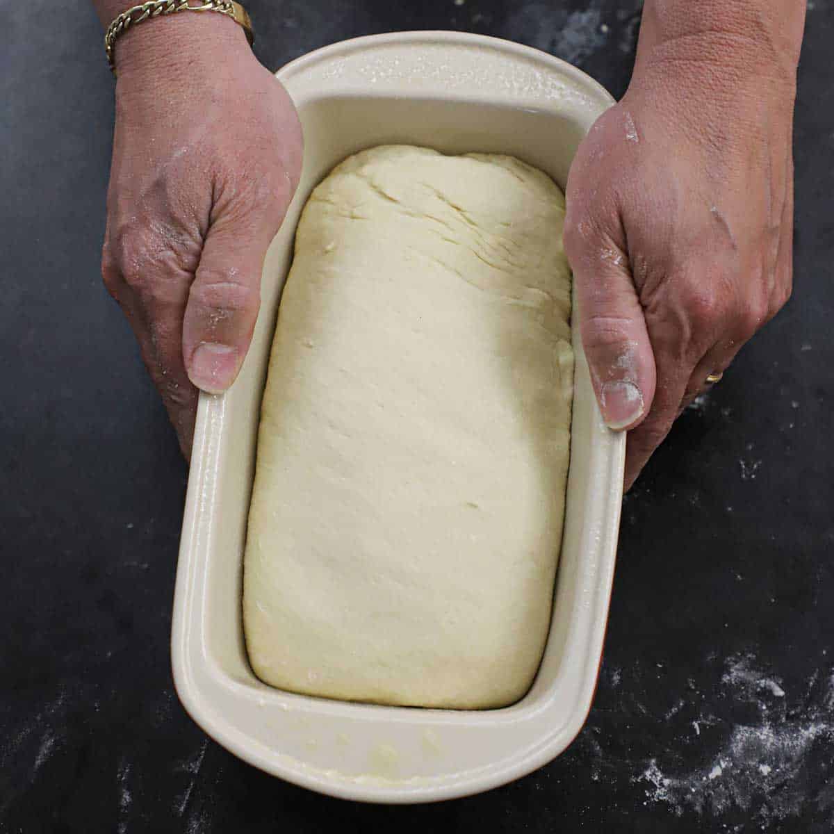 A person holding a ceramic bread loaf pan that is filled with unbaked white bread dough.