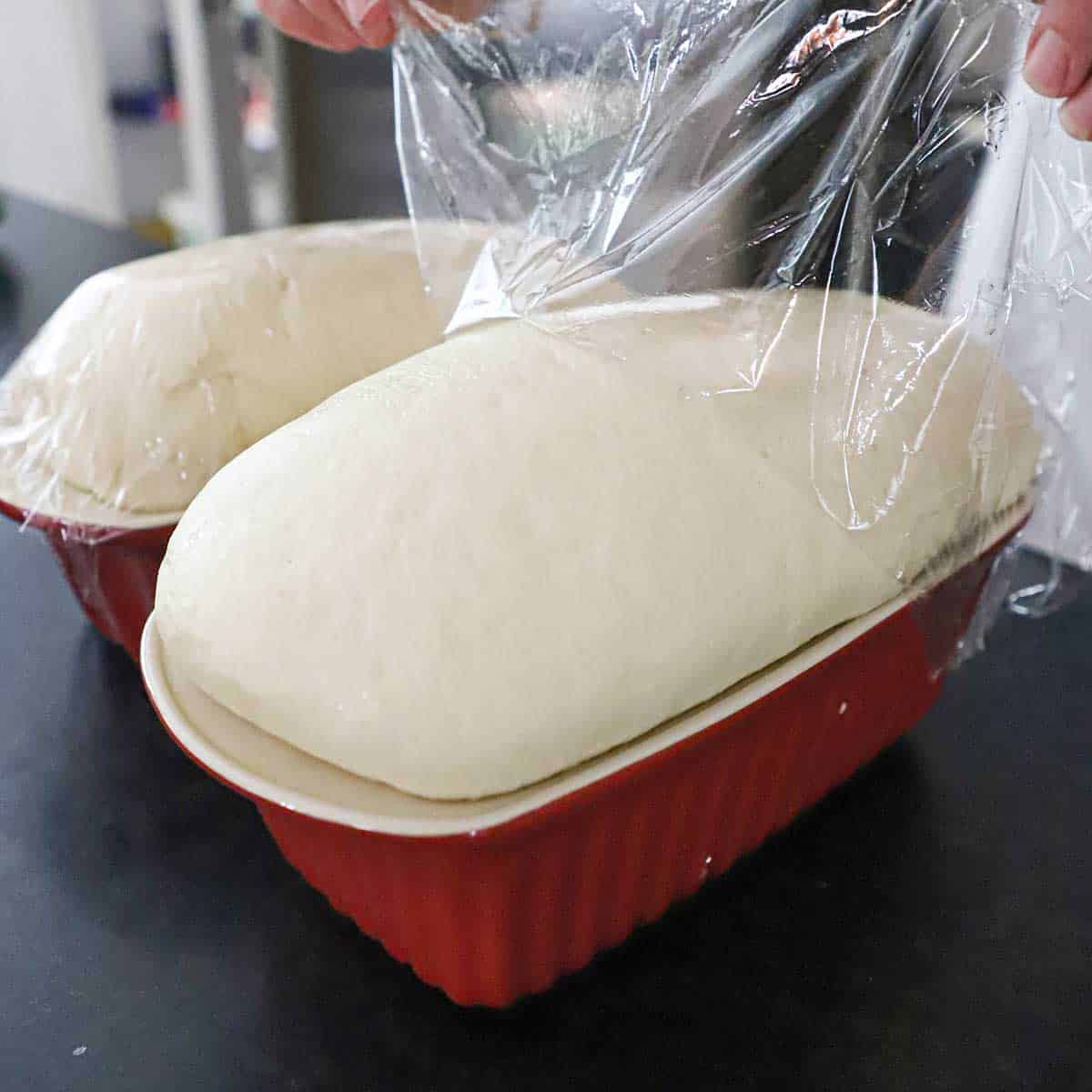 A person removing plastic wrap from the top of a fully risen loaf of country white bread in a ceramic loaf pan.