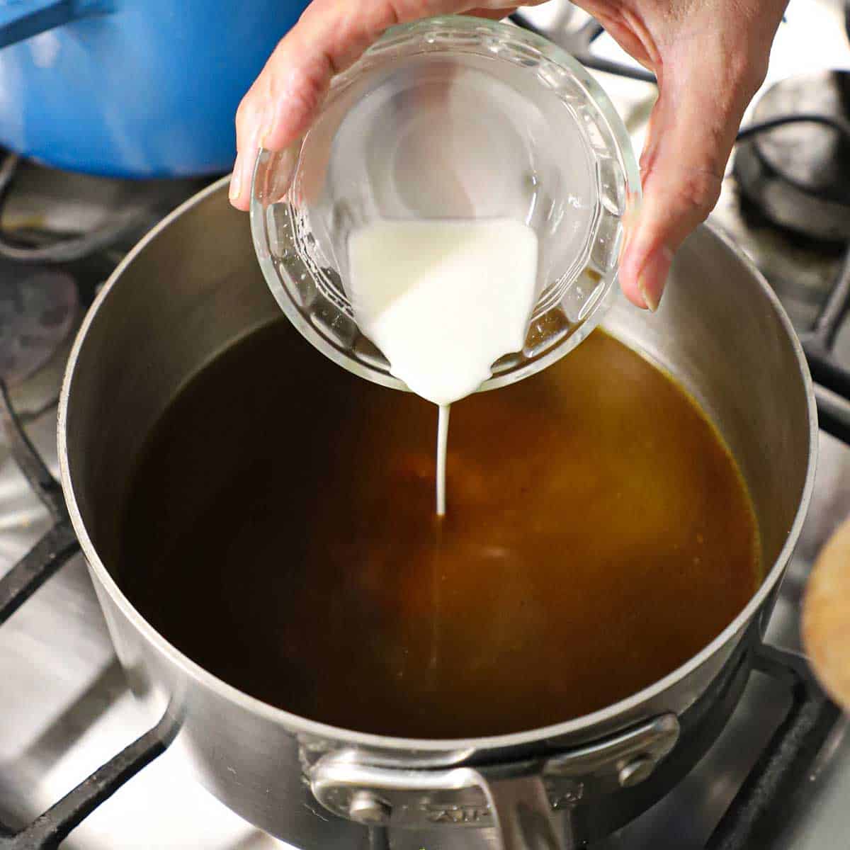 A person pouring a cornstarch slurry into a saucepan filled with simmering beef broth in it.