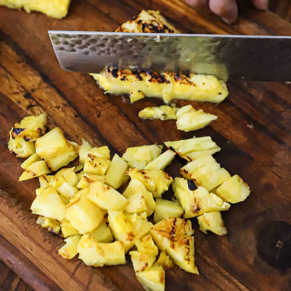 A person using a large knife to chop grilled pineapple on a wooden cutting board.