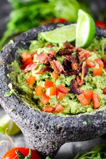 Best-ever guacamole in a black molcajete and topped with chopped tomatoes, bacon, and lime wedges.