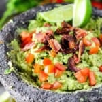 Best-ever guacamole in a black molcajete and topped with chopped tomatoes, bacon, and lime wedges.