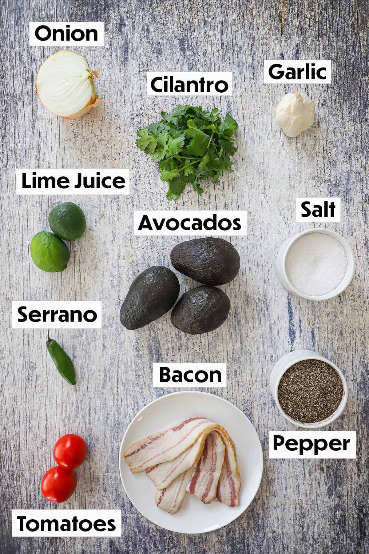 An arrangement of ingredients for guacamole on a grey wooden background including avocados, onion, cilantro, garlic, limes, pepper, bacon, tomatoes, salt, and pepper.