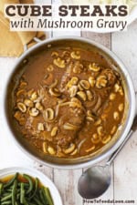 A large stainless steel skillet filled with two cube steaks with mushroom gravy and a bowl of cooked green beans and a bowl of mashed potatoes nearby.