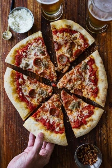A person pulling a slice of homemade New York-style pizza from the entire pie that has been cut into six slices with a small bowl of grated cheese and two glasses of beer nearby.