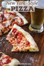 A partially eaten slice of New York-style pizza on a wooden cutting board next to more slices and a glass of beer.