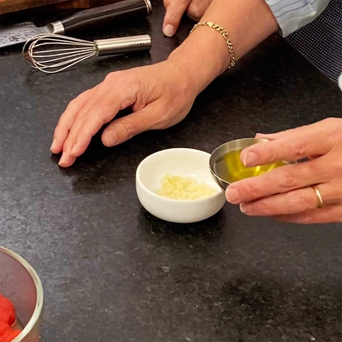 A person holding a small silver bowl filled with olive oil next to a small white bowl filled with minced garlic.