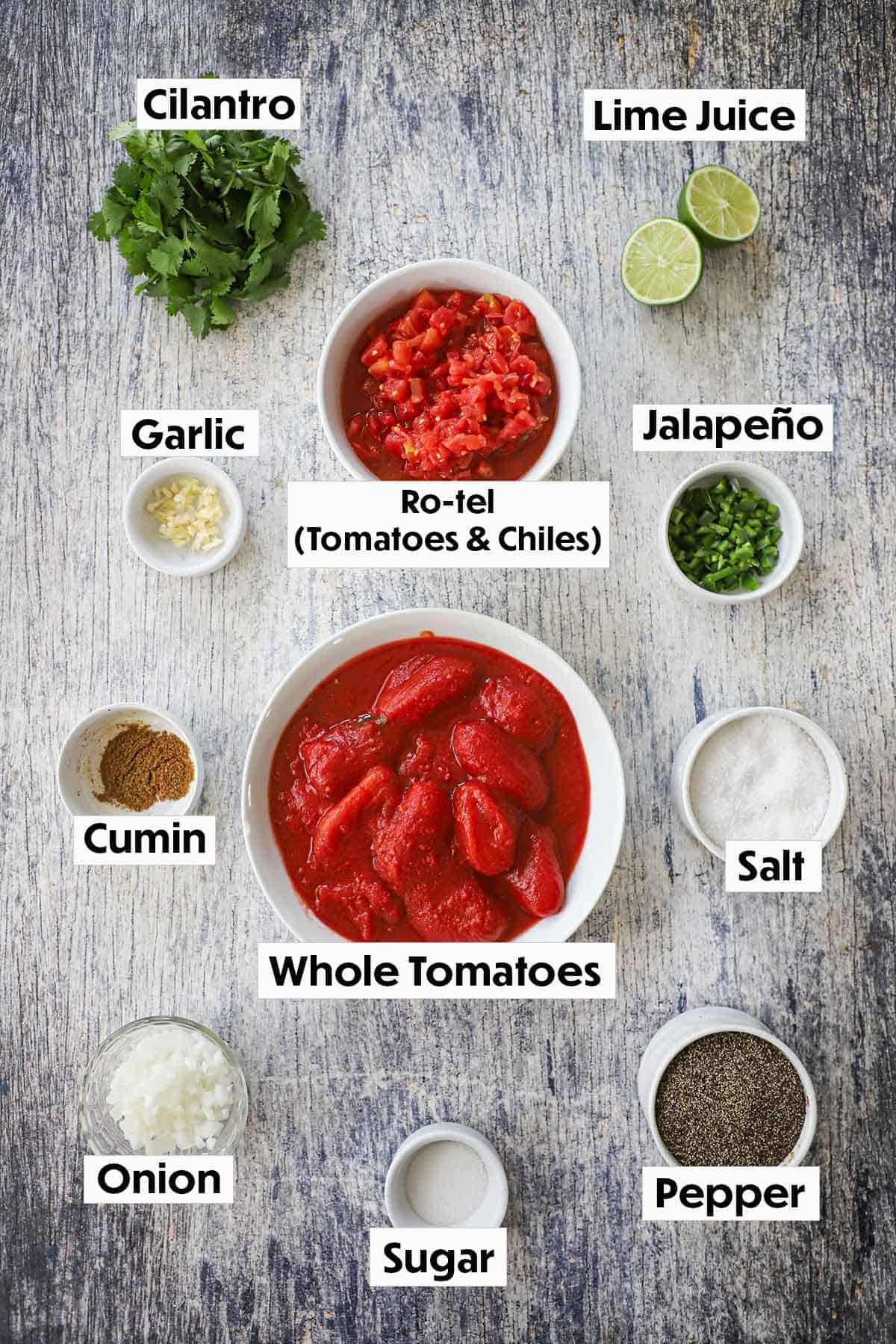 An arrangement of ingredients for restaurant-style salsa on a grey wooden background including bowls of whole tomatoes in juice, Ro-tel, limes, cilantro, garlic, onion, jalapeño, and seasonings.