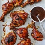 Grilled BBQ Chicken with visible grill marks resting on a metal sheet pan with a small white bowl of homemade BBQ sauce with a spoon in it nearby.
