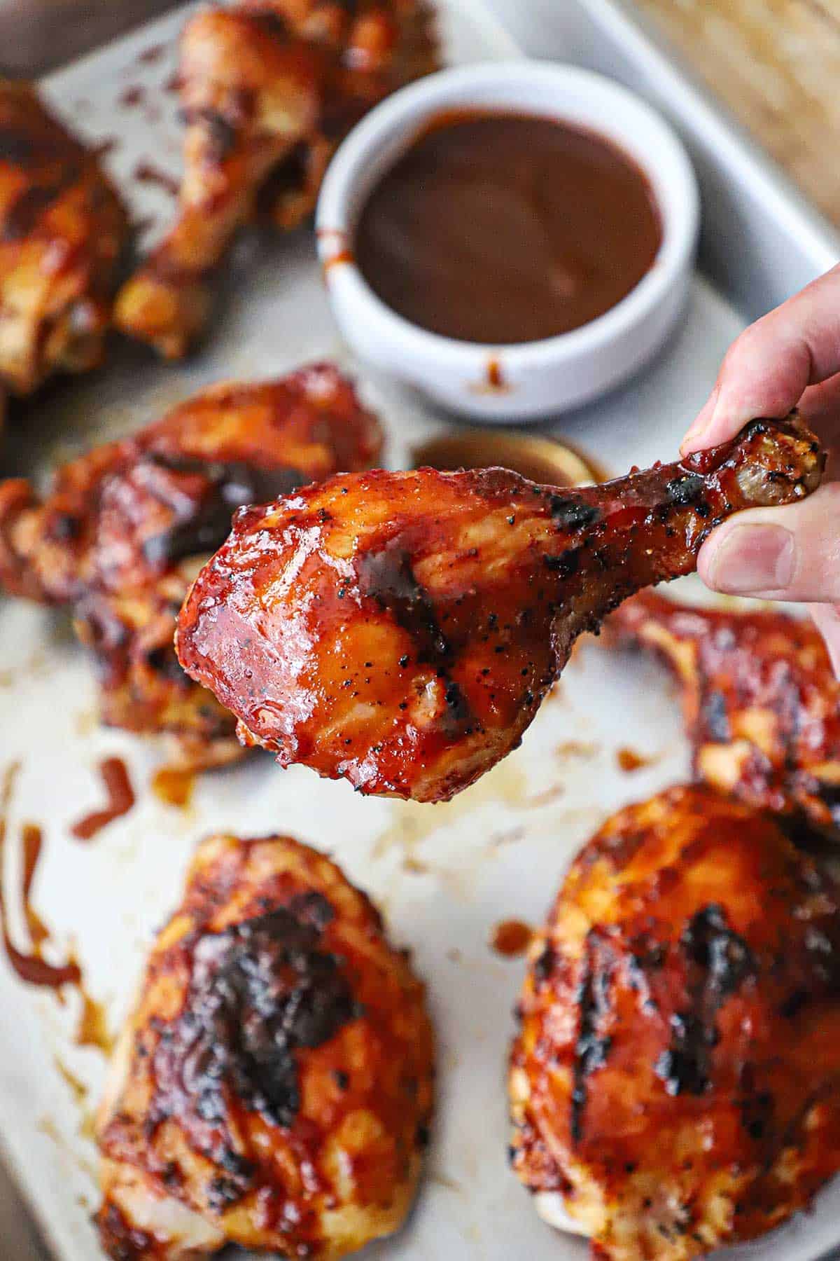A person holding a grilled BBQ chicken leg over a silver sheet pan filled with more BBQ chicken pieces and a bowl of BBQ sauce.