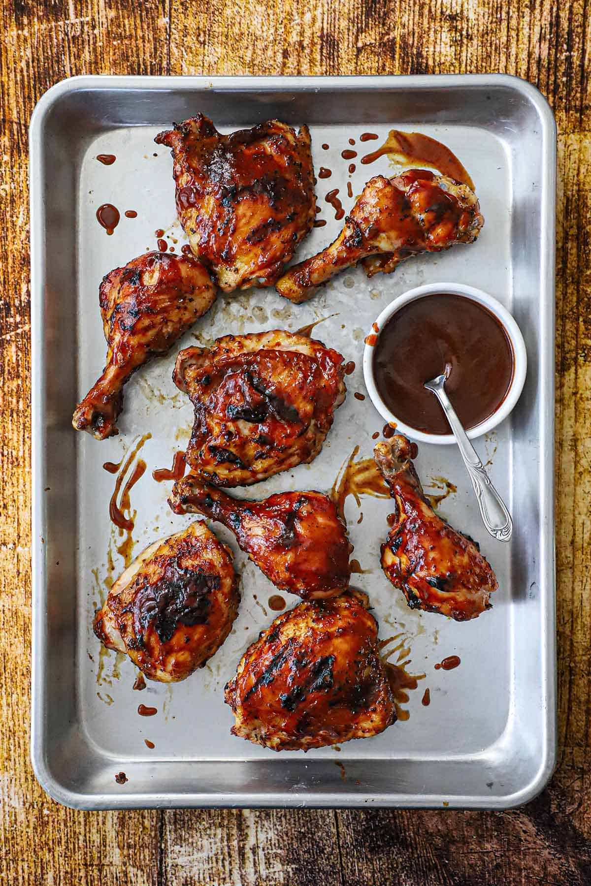 Grilled BBQ Chicken with visible grill marks resting on a metal sheet pan with a small white bowl of homemade BBQ sauce with a spoon in it nearby.