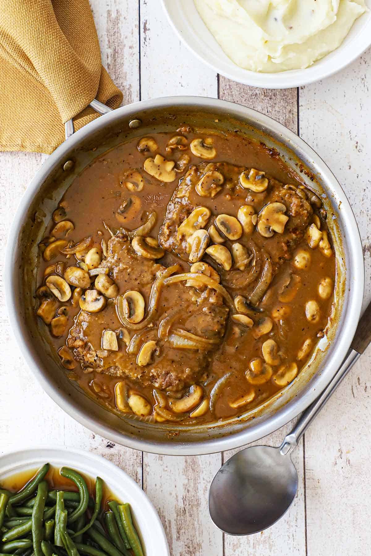 A large stainless steel skillet filled with two cube steaks with mushroom gravy and a bowl of cooked green beans and a bowl of mashed potatoes nearby.