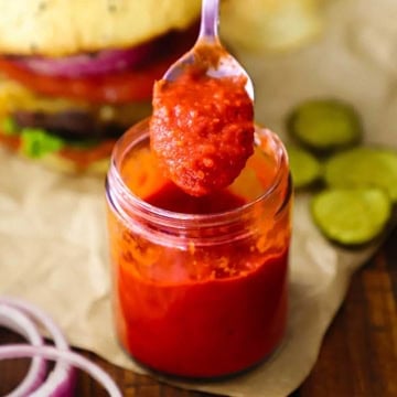 A small spoon being used to raise a spoonful of easy homemade ketchup from a small glass jar filled with the same.