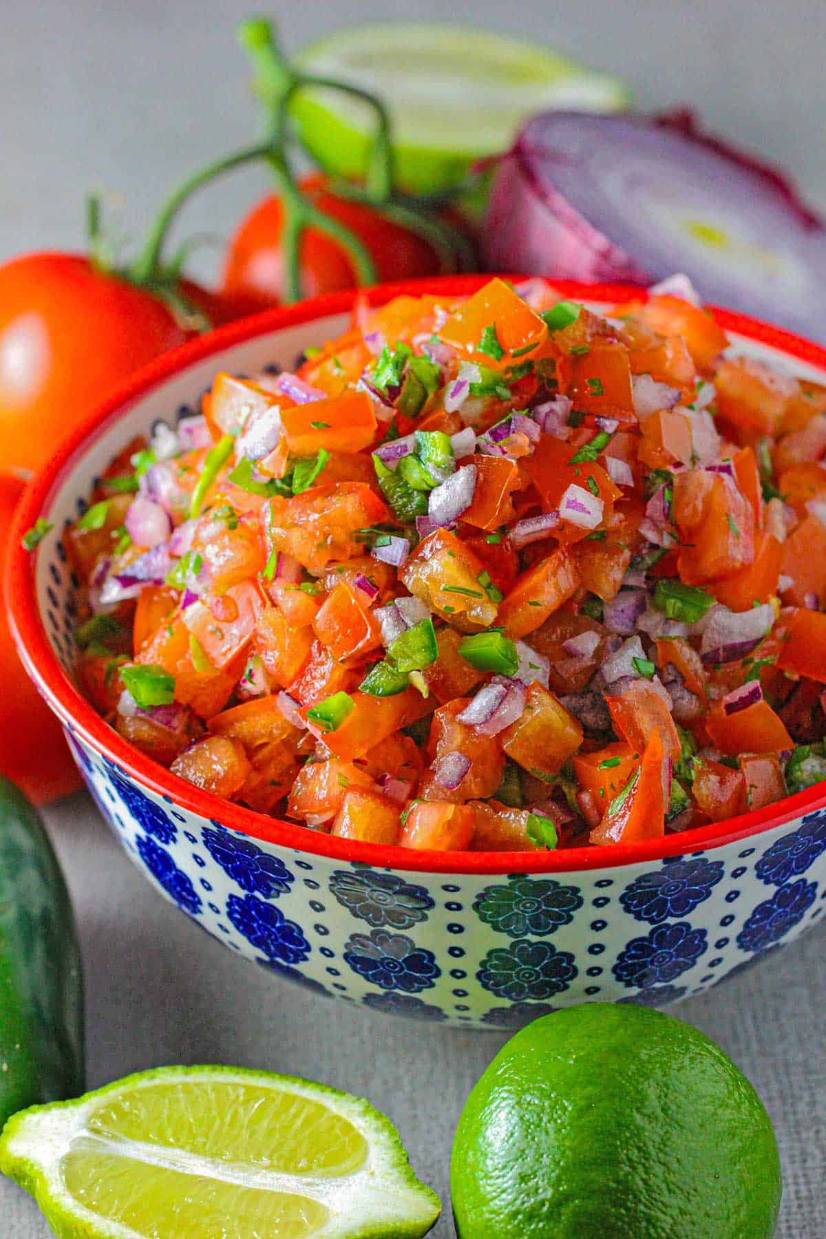 A colorful serving bowl filled with freshly made pico de gallo with limes, peppers, tomatoes, and red onion surrounding the bowl.