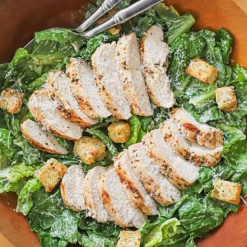 Grilled chicken Caesar salad in a large wooden bowl with homemade croutons scattered over the dressed lettuce and sliced chicken.