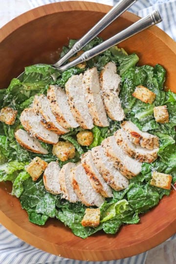 Grilled chicken Caesar salad in a large wooden bowl with homemade croutons scattered over the dressed lettuce and sliced chicken.
