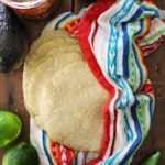 A spread-out stack of homemade corn tortillas that are nestled into a colorful napkin sitting on a wooden background and surround by whole limes, a jalapeño peppers, and an avocado.