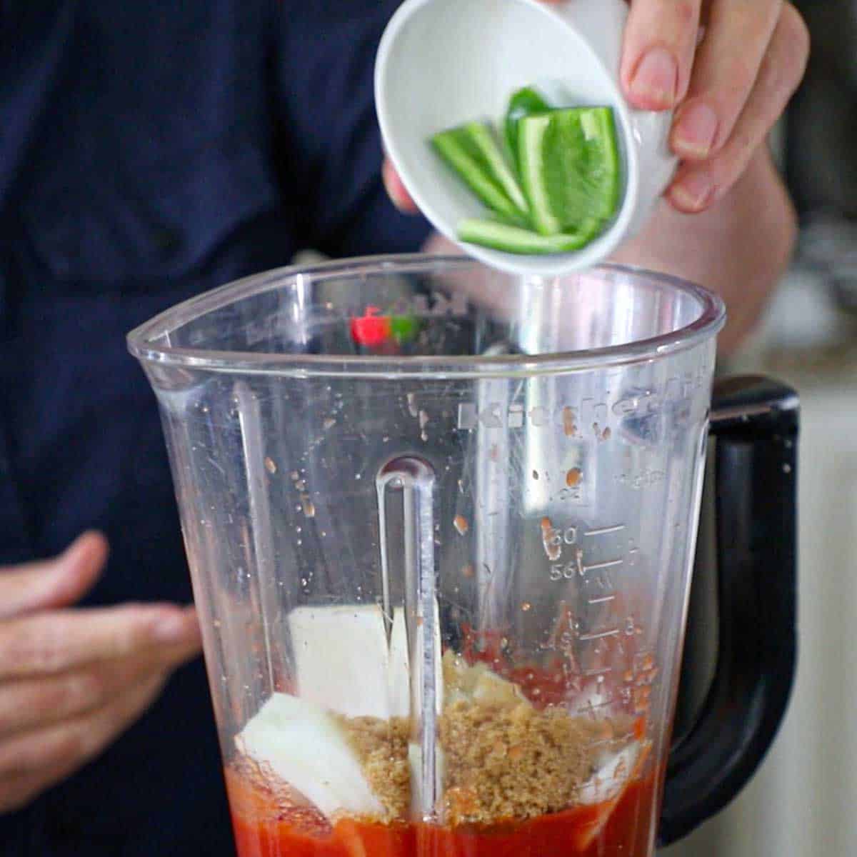 A person dumping a roughly chopped jalapeño pepper into a blender that is filled with puréed tomatoes, chopped onions, minced garlic, and light brown sugar.