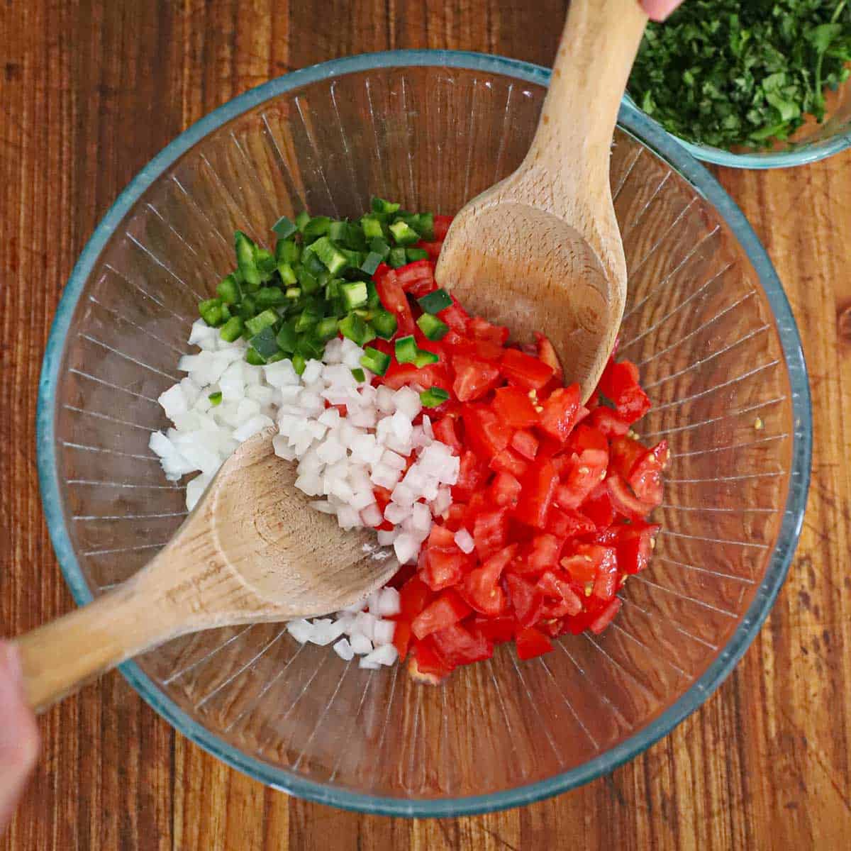 A glass bowl filled with chopped tomatoes, onion, and jalapeño with two wooden spoons tucked under the veggies.
