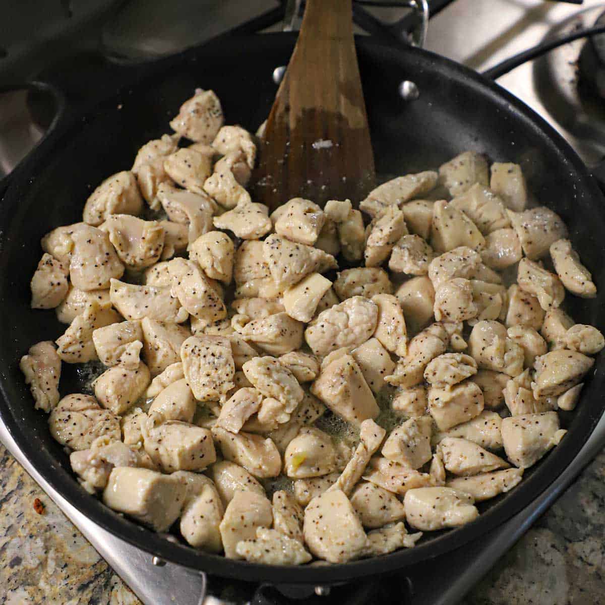 Chunks of seasoned chicken breast being sautéed in vegetable oil in a large black skillet with a wooden spatula off to the side of the pan.